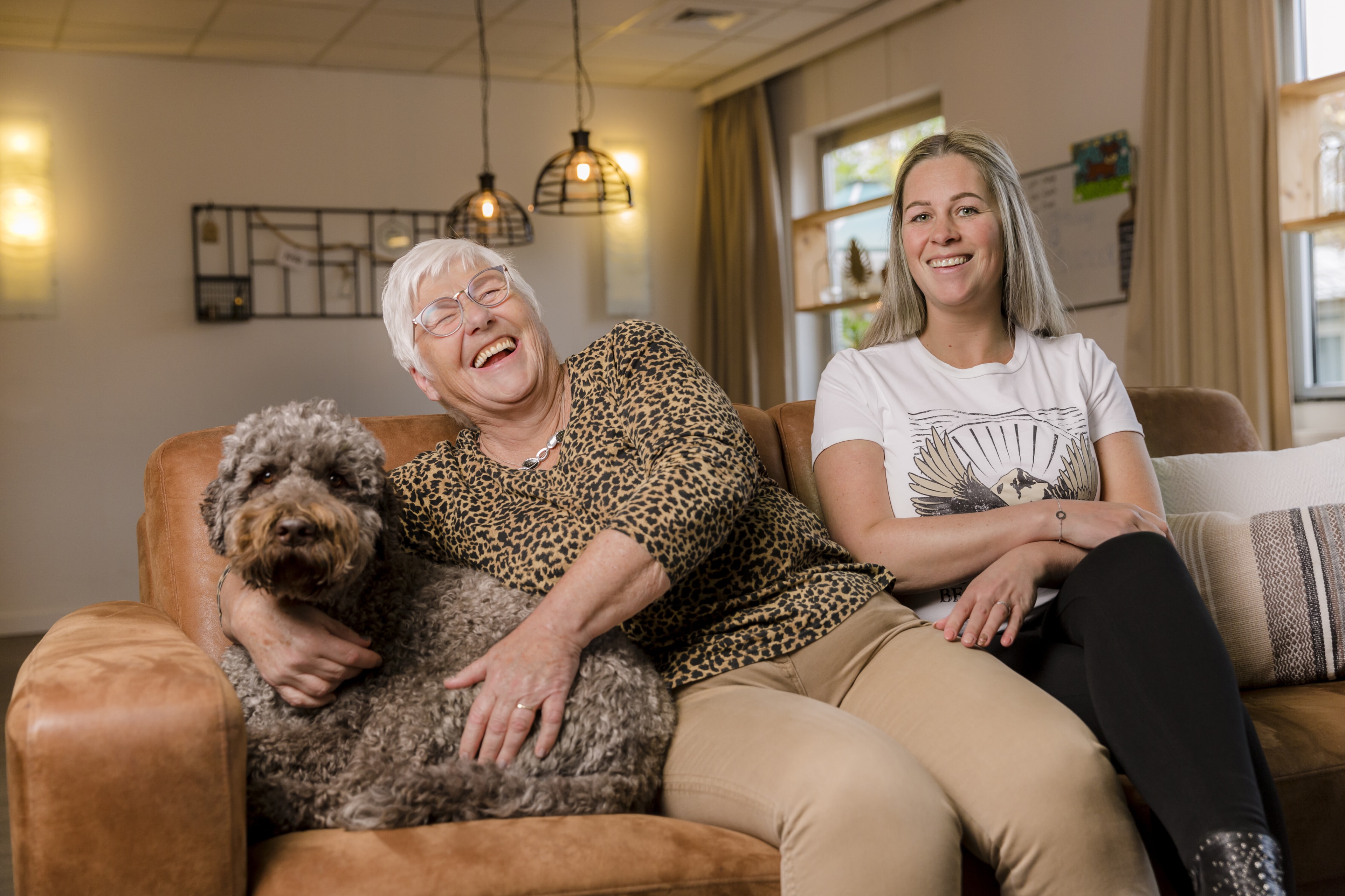 Een oudere en een jongere vrouw zitten op een bank. De ouduere vrouw lacht en naast haar zit een hond.