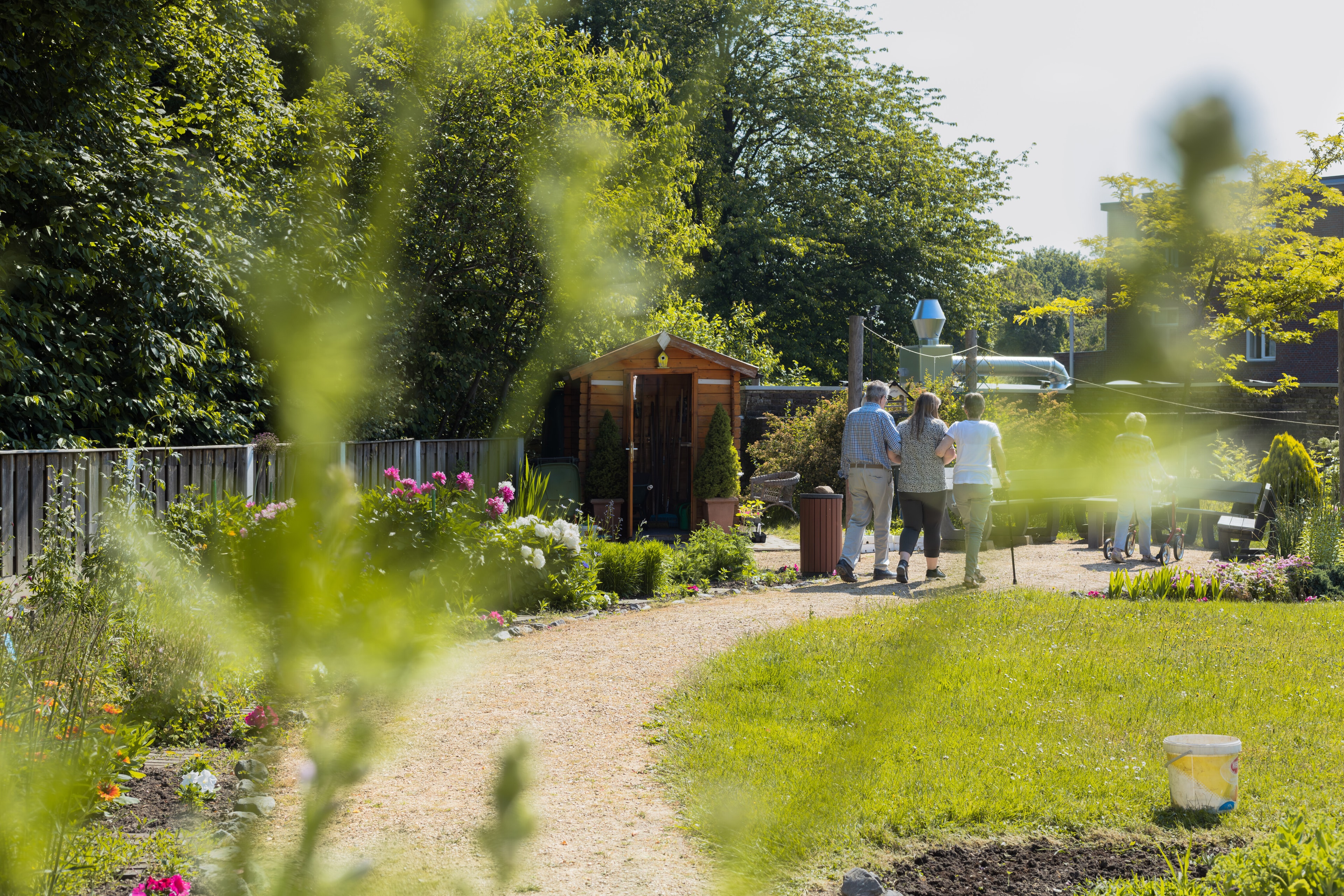 Drie personen wandelen gearmd over een paadje in een groene tuin.