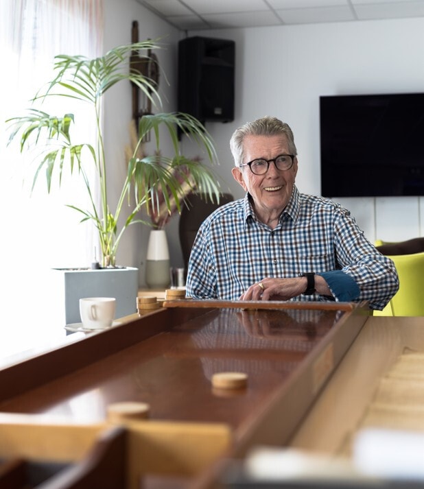 Lachende oude man met bril is aan het sjoelen. Naast de sjoelbak staat een kopje koffie.